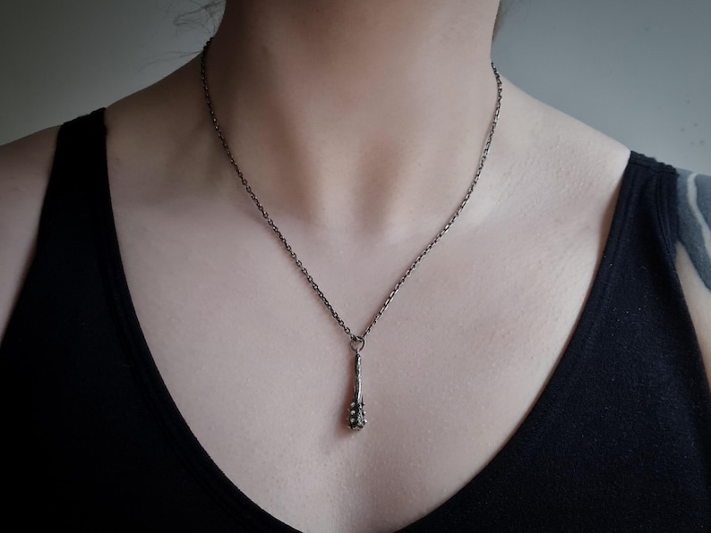 Puede incluir: Un collar de metal oscuro con un colgante delgado y texturizado en forma de peque&ntilde;a maza o garrote. El colgante cuelga de una cadena fina de metal oscuro a juego que descansa sobre el cuello, mostrando un acabado oxidado uniforme en toda la pieza.