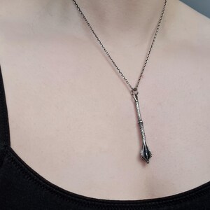 May include: A silver-toned metal pendant shaped like a medieval mace, featuring intricate textured detailing on the handle and a flanged head. The pendant hangs from a thin, dark silver-colored chain that rests against the chest area.
