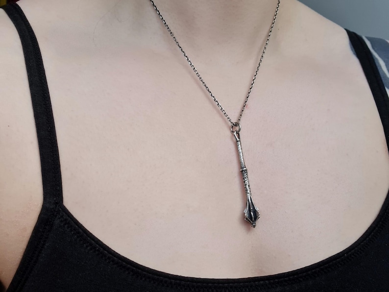 May include: A silver-toned metal pendant shaped like a medieval mace, featuring intricate textured detailing on the handle and a flanged head. The pendant hangs from a thin, dark silver-colored chain that rests against the chest area.