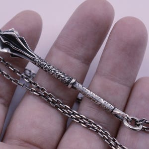 May include: A silver pendant shaped like a medieval mace with a textured handle and a multi-faceted head, attached to a silver chain. The metal has a dark, oxidized finish in the recessed areas, highlighting the intricate details of the weapon design.