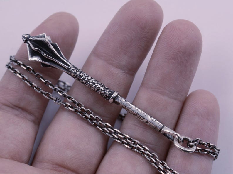 May include: A silver pendant shaped like a medieval mace with a textured handle and a multi-faceted head, attached to a silver chain. The metal has a dark, oxidized finish in the recessed areas, highlighting the intricate details of the weapon design.
