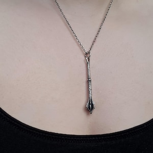 May include: A silver-toned metal pendant shaped like a mace, suspended from a delicate matching silver-toned chain. The pendant features a textured handle and a pointed, multi-faceted head, hanging vertically against a plain black fabric background.