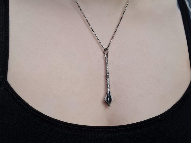 May include: A silver-toned metal pendant shaped like a mace, suspended from a delicate matching silver-toned chain. The pendant features a textured handle and a pointed, multi-faceted head, hanging vertically against a plain black fabric background.