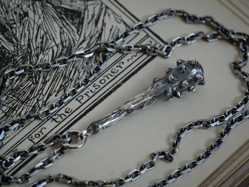 May include: A silver-toned metal pendant shaped like a spiked club or mace, featuring a textured, weathered surface and small protruding spikes at the top. The pendant is attached to a thick, interlocking silver-toned metal chain necklace.