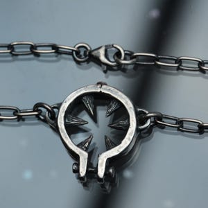 Puede incluir: Un collar de cadena de plata con un colgante circular que presenta un diseño de estrella con bordes puntiagudos.