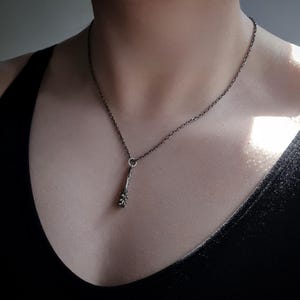 Puede incluir: Un collar de tono plateado oscuro con un colgante delgado y texturizado en forma de peque&ntilde;a maza o rama r&uacute;stica. El colgante cuelga de una delicada cadena de metal oscuro a juego que descansa sobre la clav&iacute;cula, mostrando un acabado met&aacute;lico.