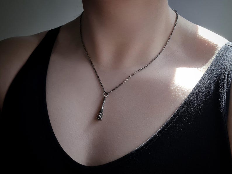 Puede incluir: Un collar de tono plateado oscuro con un colgante delgado y texturizado en forma de peque&ntilde;a maza o rama r&uacute;stica. El colgante cuelga de una delicada cadena de metal oscuro a juego que descansa sobre la clav&iacute;cula, mostrando un acabado met&aacute;lico.