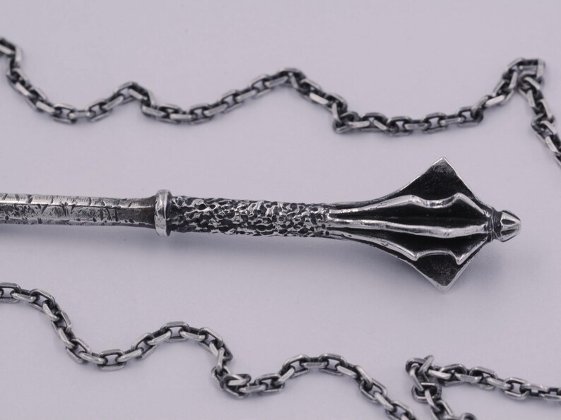 May include: A silver-toned metal pendant shaped like a medieval mace with a textured handle and a flanged head, suspended from a matching silver-toned link chain. The metal features a dark, oxidized finish that highlights the intricate details.