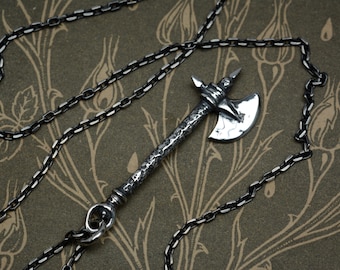 May include: A silver-toned pendant shaped like a battle axe with a textured, hammered handle and a smooth, curved blade. The pendant is attached to a dark, oxidized silver chain with rectangular links, creating a cohesive metallic accessory.