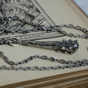 Puede incluir: Un collar de plata con un colgante en forma de maza medieval, que presenta un mango texturizado de tonos oscuros y una cabeza con p&uacute;as. El colgante est&aacute; unido a una cadena de eslabones de plata a juego, con un acabado met&aacute;lico detallado y envejecido.