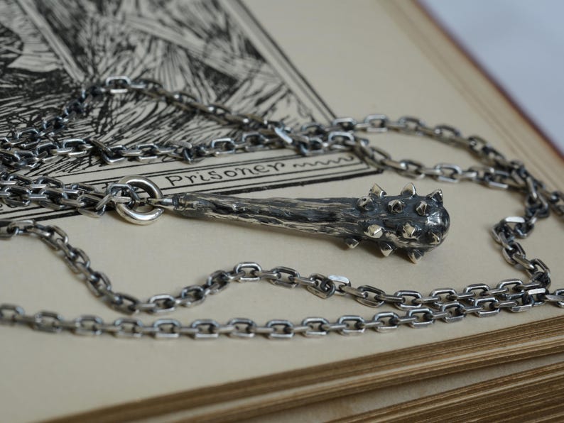 Puede incluir: Un collar de plata con un colgante en forma de maza medieval, que presenta un mango texturizado de tonos oscuros y una cabeza con p&uacute;as. El colgante est&aacute; unido a una cadena de eslabones de plata a juego, con un acabado met&aacute;lico detallado y envejecido.