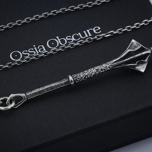 May include: A silver-toned pendant shaped like a medieval mace with a textured handle and a multi-faceted head, suspended from a matching silver-toned chain. The pendant rests on a black box featuring the white text Ossia Obscure printed on it.