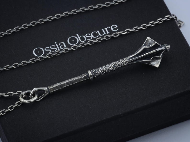 May include: A silver-toned pendant shaped like a medieval mace with a textured handle and a multi-faceted head, suspended from a matching silver-toned chain. The pendant rests on a black box featuring the white text Ossia Obscure printed on it.