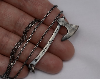 May include: A silver-toned metal pendant shaped like a small battle axe with a textured, weathered finish. The handle of the axe features engraved runic symbols. The pendant is attached to a thin, dark silver-colored metal chain link necklace.