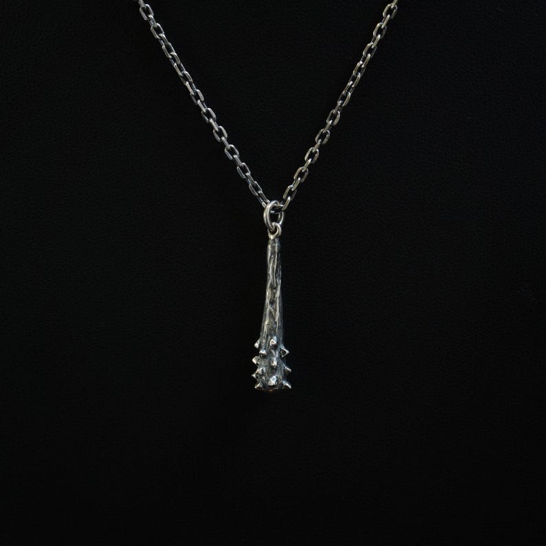 Puede incluir: Un collar de tono plateado con un colgante en forma de maza texturizada con p&uacute;as. El colgante cuelga de una delicada cadena de eslabones a juego. La superficie met&aacute;lica tiene un acabado oscuro y oxidado que resalta los detalles rugosos.