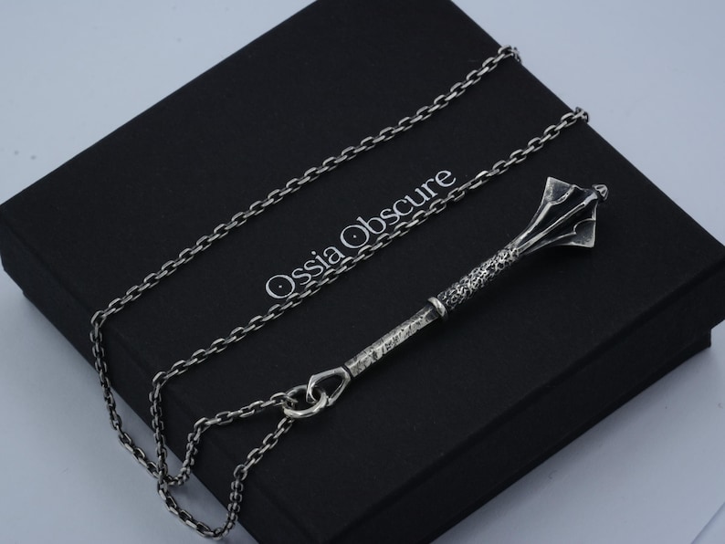 May include: A silver-toned metal pendant shaped like a medieval mace, featuring a textured handle and a flanged head, suspended from a matching silver-toned link chain. The jewelry rests on a black box with the text Ossia Obscure printed in white.