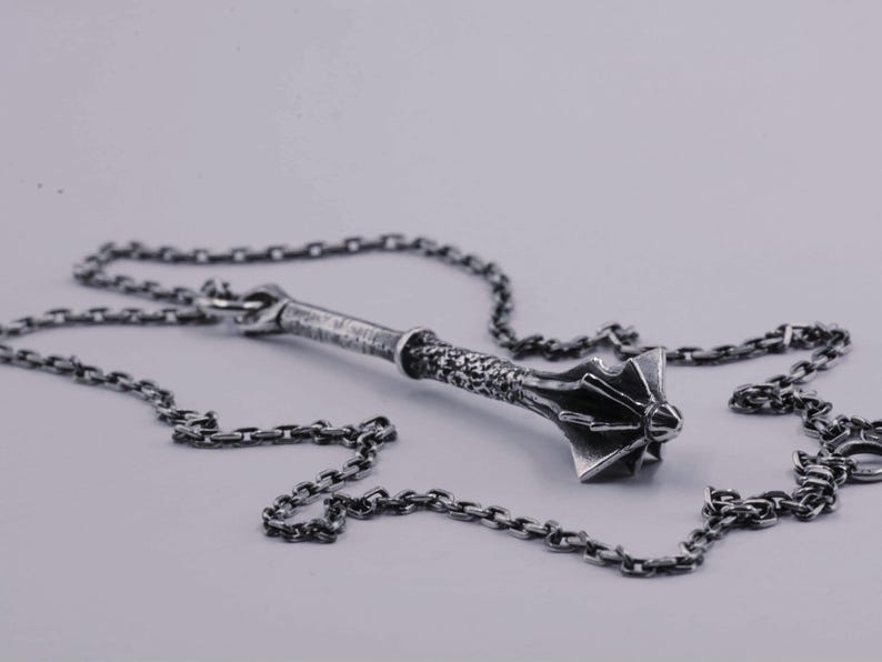 May include: A silver-toned metal pendant shaped like a medieval mace, featuring a textured handle and a flanged head, suspended from a matching silver-toned link chain. The metal has a darkened, oxidized finish that highlights the intricate details.
