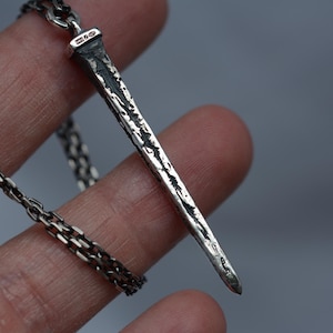 May include: A silver chain necklace with a silver nail pendant. The nail is textured and has a hammered finish.