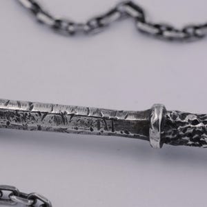 May include: A silver-toned metal pendant featuring a textured, weathered surface with deep grooves and a rough, hammered finish, attached to a matching silver-toned link chain that loops across the top and bottom of the frame.