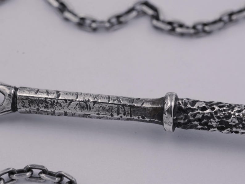 May include: A silver-toned metal pendant featuring a textured, weathered surface with deep grooves and a rough, hammered finish, attached to a matching silver-toned link chain that loops across the top and bottom of the frame.