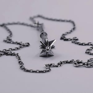 May include: A silver-toned metal pendant shaped like a spiked mace head, featuring a textured handle and a pointed star-like top. The pendant is attached to a matching silver-toned metal link chain that rests in a circular pattern around the object.