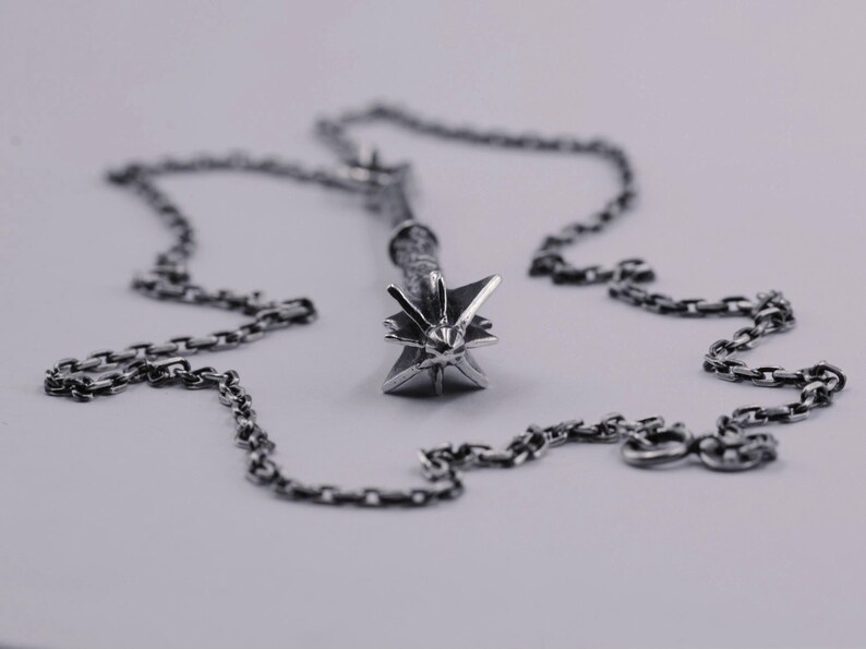 May include: A silver-toned metal pendant shaped like a spiked mace head, featuring a textured handle and a pointed star-like top. The pendant is attached to a matching silver-toned metal link chain that rests in a circular pattern around the object.