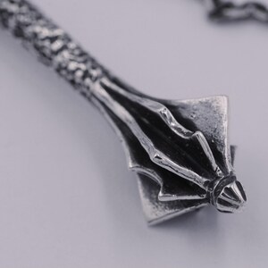 May include: A silver-toned metal pendant shaped like a mace with a textured, cylindrical handle and a multi-flanged head. The piece features an oxidized finish that highlights the intricate details of the metalwork and is attached to a chain.