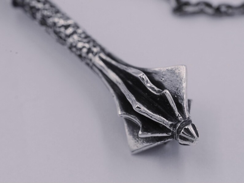May include: A silver-toned metal pendant shaped like a mace with a textured, cylindrical handle and a multi-flanged head. The piece features an oxidized finish that highlights the intricate details of the metalwork and is attached to a chain.