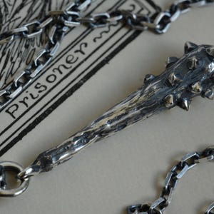 Puede incluir: Un colgante de tono plateado con forma de maza con p&uacute;as, que presenta una superficie texturizada y desgastada con una serie de peque&ntilde;as protuberancias puntiagudas en la cabeza. El colgante est&aacute; unido a un collar de cadena de eslabones gruesos.