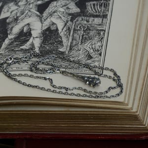 Puede incluir: Un collar de metal plateado con un colgante en forma de maza con p&uacute;as. La cadena est&aacute; compuesta por peque&ntilde;os eslabones ovalados entrelazados. El colgante tiene un acabado oscuro y texturizado con una cabeza redondeada y nudosa.