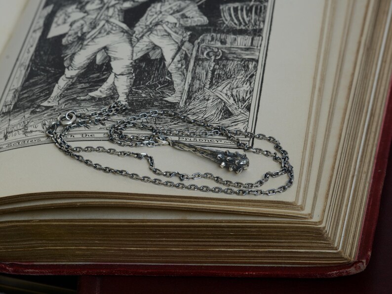 Puede incluir: Un collar de metal plateado con un colgante en forma de maza con p&uacute;as. La cadena est&aacute; compuesta por peque&ntilde;os eslabones ovalados entrelazados. El colgante tiene un acabado oscuro y texturizado con una cabeza redondeada y nudosa.