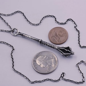 May include: A silver-toned metal mace pendant on a matching link chain, displayed alongside a U.S. quarter coin for scale. The pendant features a textured handle and a multi-flanged head, showcasing a detailed, weathered metallic finish throughout.