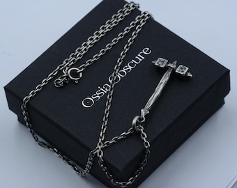 May include: A silver-toned necklace featuring a pendant shaped like a double-headed hammer with a textured handle. The pendant is attached to a delicate link chain. The jewelry is displayed on a black box with the text Ossia Obscure printed in white.