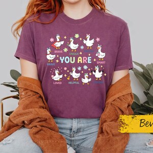 Può includere: Una t-shirt color prugna con una grafica raffigurante diverse anatre bianche in stile cartoon disposte in cerchio. Il testo "YOU ARE" è centrato al centro, circondato da parole come "BRAVE", "UNIQUE", "SMART", "LOVED", "HELPFUL" e "STRONG".