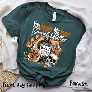 May include: Forest green t-shirt with the text "You Inspire My Inner Serial Killer." The graphic includes a poison bottle, skull, and orange roses. The shirt is made of a soft material.