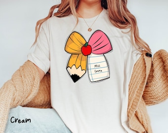 Puede incluir: Camiseta color crema con un diseño de lápiz y lazo con un acento de manzana roja. El diseño incluye el texto "Miss Emma" en un bloc de notas. La camiseta es de estilo casual.