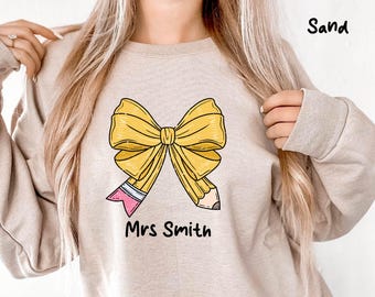 Puede incluir: Sudadera color arena con un lazo amarillo hecho de lápices. Los lápices tienen gomas de borrar rosas y puntas grises. El texto "Mrs Smith" está impreso debajo del lazo en negro. La sudadera tiene mangas largas.