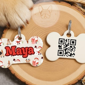 Puede incluir: Dos etiquetas para mascotas en forma de hueso. Una etiqueta es de color crema con el nombre "Maya" en rojo, rodeada de ilustraciones navideñas. La otra etiqueta es blanca con un código QR. Ambas etiquetas tienen llaveros plateados.