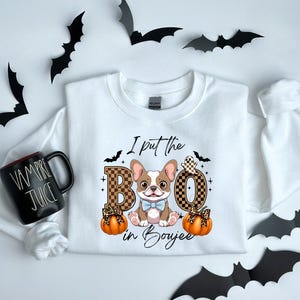 Puede incluir: Sudadera blanca con un gráfico de un bulldog francés con pajarita azul, calabazas y el texto "I put the BOO in Boujee". Murciélagos negros decoran el fondo. Una taza negra con "Vampire Juice" está a la izquierda.