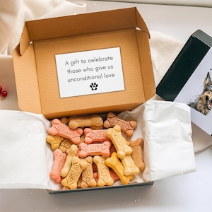 Puede incluir: Una caja de cartón abierta llena de golosinas para perros en forma de hueso en tonos rosa, beige y amarillo. Una tarjeta blanca dentro de la caja dice: "Un regalo para celebrar a quienes nos dan amor incondicional" con una huella de pata. Una caja negra con una ilustración de perro.