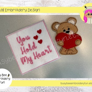 May include: Embroidered design featuring a brown teddy bear holding a red heart and a white square with the text "You Hold My Heart" in pink thread.