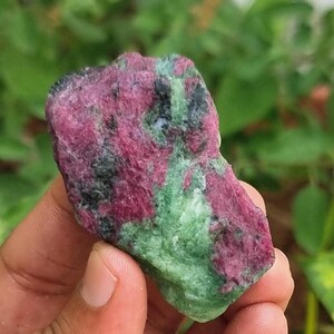 May include: A natural gemstone with a blend of colours. The stone displays a deep red, with areas of black and green. The texture appears rough and natural, with visible mineral formations. The gemstone is held in a hand.