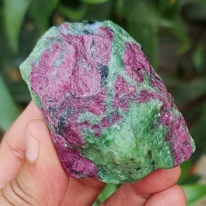Pode incluir: Uma pedra bruta de rubi e zoisite, segurada por uma mão. A pedra tem uma aparência manchada com áreas de rosa profundo e verde. As cores são naturais e a textura é áspera e irregular. É um espécime mineral natural.