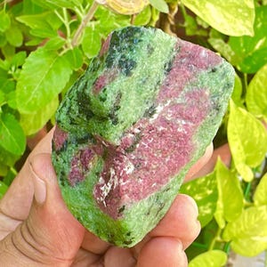 May include: A natural stone with a mix of colours. The stone has a green base with streaks of deep pink and black. The stone is held in a hand, with green foliage in the background.
