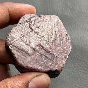 Puede incluir: Un gran cristal de rubí rosa áspero con una superficie estriada. El cristal está facetado y tiene una forma hexagonal.