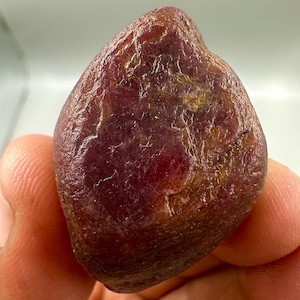 Könnte beinhalten: Ein roher, ungeschliffener Rubin-Edelstein mit tiefroter Farbe und einer leicht unregelmäßigen Form. Die Oberfläche hat ein strukturiertes Aussehen mit Farbvariationen, einschließlich dunklerer und heller Bereiche. Der Edelstein wird in einer Hand gehalten.