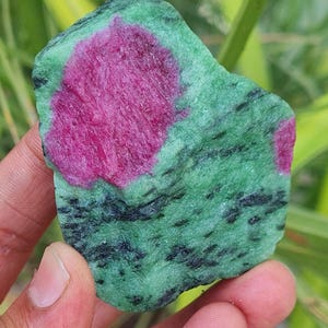 Puede incluir: Una gema verde y rosa en bruto con un patrón moteado. La gema tiene una gran área rosa y áreas rosas más pequeñas dispersas.