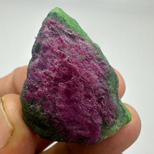 Peut inclure: Une pierre précieuse brute avec un cœur magenta vif et une couche extérieure verte. La surface de la pierre présente un aspect texturé, mettant en valeur les formations minérales naturelles. Le bijou est tenu dans une main, soulignant sa taille et sa forme.