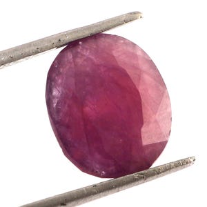 Może przedstawiać: Fasetowany, owalny kamień szlachetny rubinu, trzymany przez metalową pęsetę. Rubin ma głęboki, nasycony różowo-czerwony kolor z widocznymi wtrąceniami wewnętrznymi. Kamień jest na białym tle.