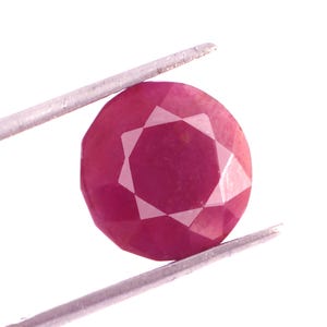 Peut inclure: Une pierre précieuse rubis ronde et facettée, tenue par une pince argentée. Le rubis est d'un rouge profond avec de multiples facettes qui réfléchissent la lumière. La pince est fine et métallique.